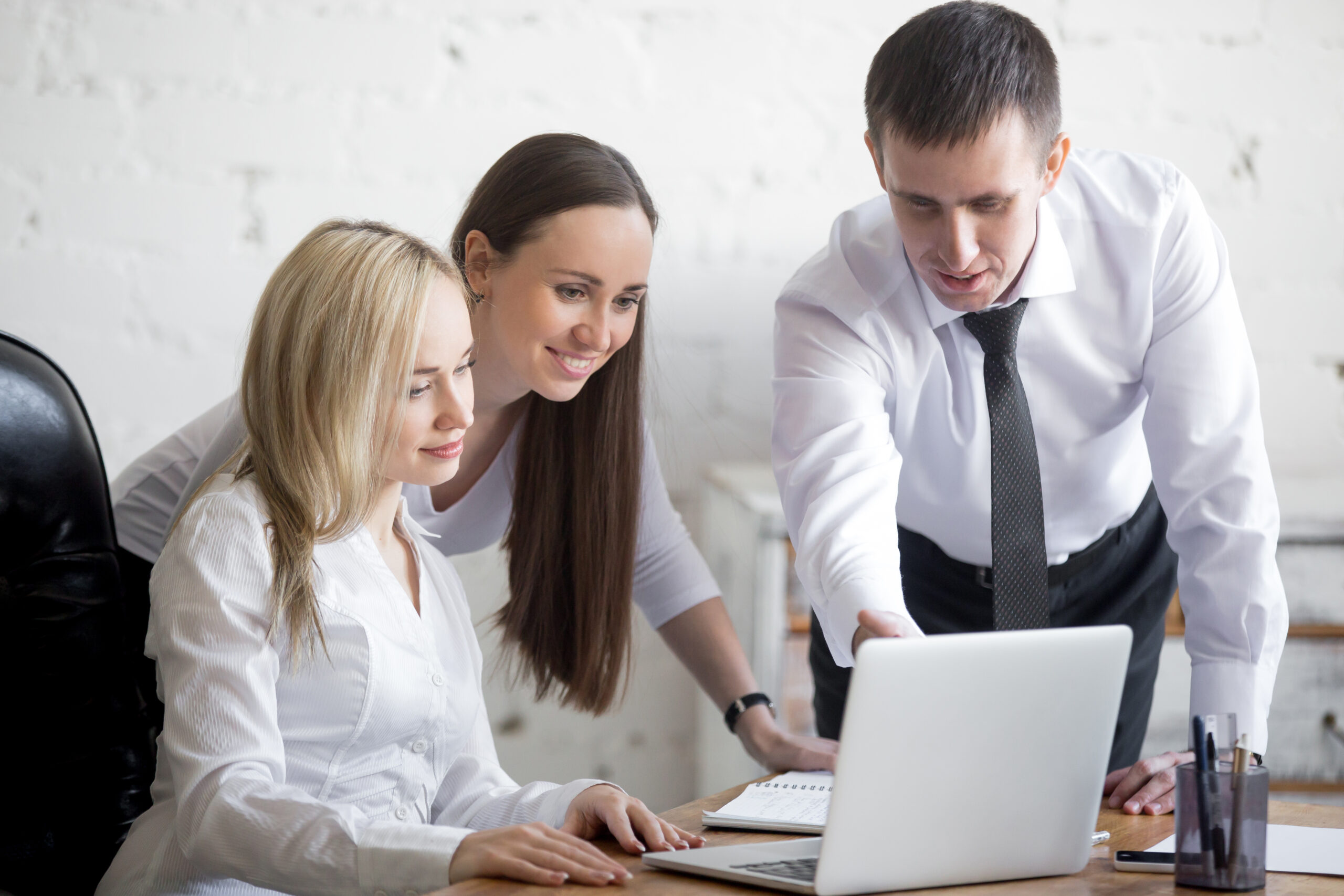 Teamwork concept. Group of three smiling young people working on project at office desk. Business team in formal wear using computer while discussing cooperative work